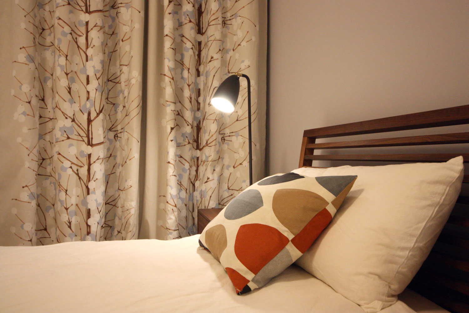 Eine schwarze Nachttischlampe beleuchtet stimmungsvoll ein farbiges Kissen auf einem Bett. Im Hintergrund ein Vorhang mit einem Baummotiv.