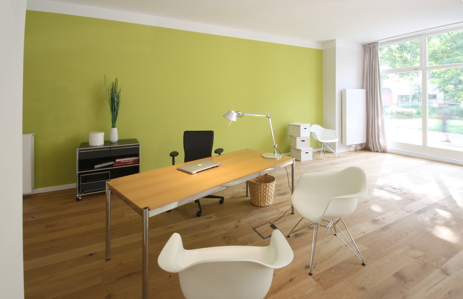 Beratungssituation in einem großen, hellen Raum. Schreibtisch mit Holzplatte und 2 weißen Schalenstühlen aus Kunststoff. Die hintere Raumwand ist hellgrün.