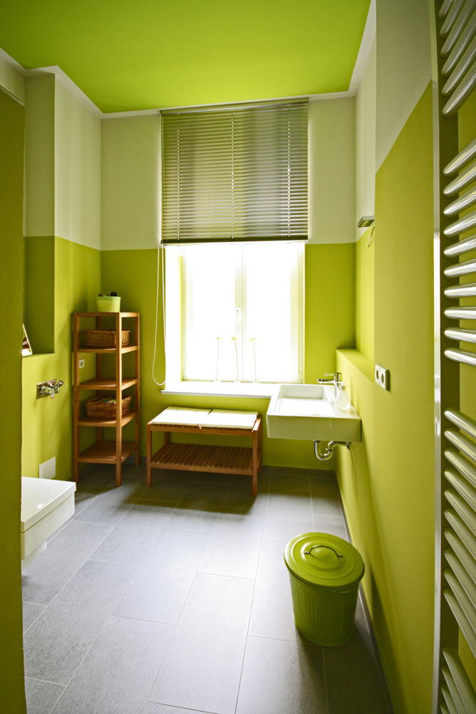 Kleines Badezimmer mit hellgrünen Wänden und hellgrünem Deckenfeld und Holzregalen. Im Vordergrund ein passender grüner Metalleimer.