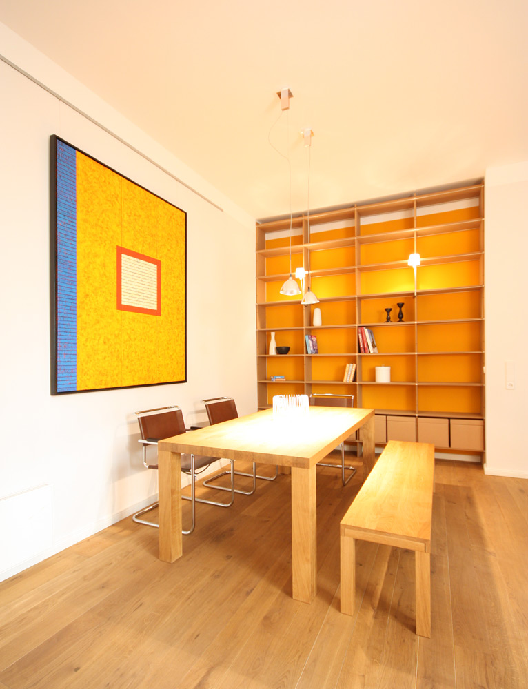 Heller Essbereich mit Tisch und Sitzbank aus hellem Holz, im Hintergrund ein raumhohes Regal aus MDF mit oranger Rückwand. Links an der Wand ein großes abstraktes Bild in gelborange, blau und rot.