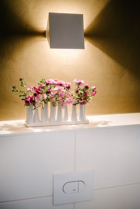 Eine klappbare weiße Wandleuchte mit indirektem Licht. Darunter eine Vase mit rosafarbenen Blumen