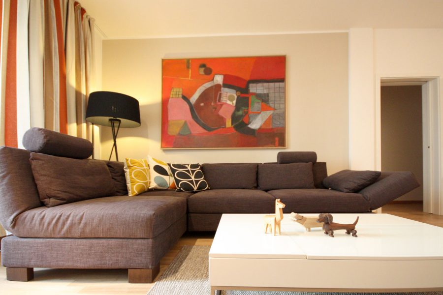 EIn gemütliches und elegantes Wohnzimmer mit einer grauen Eckcouch und einem weißen quadratischen Sofatisch. An der Wand ein warmgraues Farbfeld und ein Gemälde in kräftigen Rot-Tönen. Ein gestreifter Vorhang in rostrot, weiß und Brauntönen.