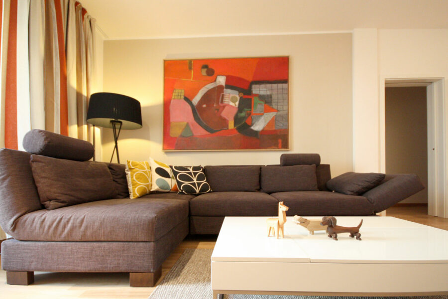 EIn gemütliches und elegantes Wohnzimmer mit einer grauen Eckcouch und einem weißen quadratischen Sofatisch. An der Wand ein warmgraues Farbfeld und ein Gemälde in kräftigen Rot-Tönen. Ein gestreifter Vorhang in rostrot, weiß und Brauntönen.
