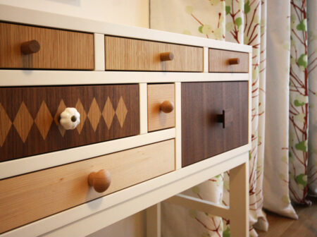 Sideboard in Weiß und mit verschiedenen Holzarten. Im Hintergrund ein Vorhang mit grünem Baummotiv.