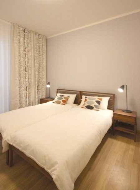 Holzbett aus Nussbaum mit weißem Bettbezug und bunten Kissen von Marimekko. Vorhänge mit einem Baumblütenmotiv und eine hellblaue Wand im Hintergrund. Schwarze Tischleuchten auf Nachttischen aus Nussbaum.