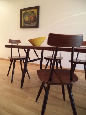 Holzstühle und Holz-Esstisch in skandinavischem Design mit gelber Porzellanschale. Im Hintergrund hängt ein Bild an der Wand, ein Stilleben mit Früchten.