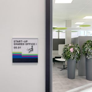 Wegeleitsystem mit farbigen Streifen auf einem Türschild; im Hintergrund ein Blick in einen Coworking-Space