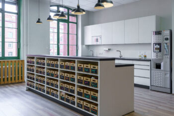 Weiße Küchenzeile mit Tresen und individuellen Mitarbeiter-Boxen in einem Industrie-Altbau.