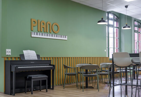 Vor einer grünen Wand mit dem Schriftzug "PIANO" und einer Klaviaturgrafik steht ein echtes schwarzes Klavier. Daneben einige Cafeteriatische.