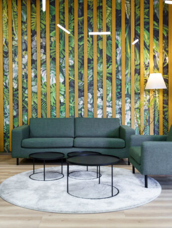 Gemütliche Sitzecke: Ein dunkelgrünes Sofa steht vor einer Wand aus Blättertapete und Eiche-Holzlamellen. Davor stehen 3 runde, schwarze Sofatische aus Metall auf einem flauschigen weißen Teppich.