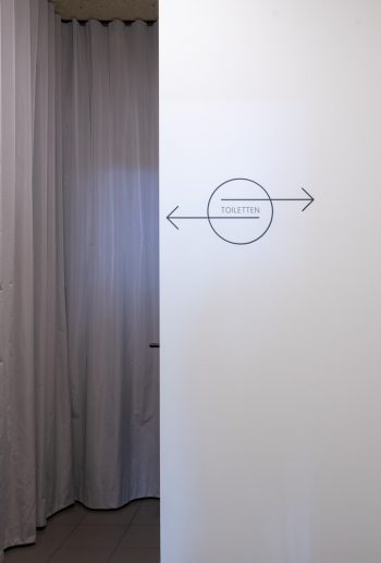 Ein grafisches Piktogramm weist zu den Toilettenräumen; im Hintergrund ein grauer Vorhang, der den Stauraum versteckt.
