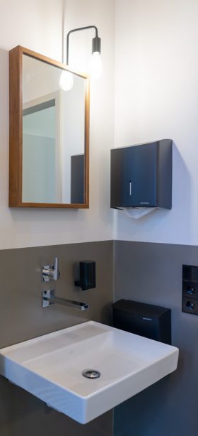 Schlichte Eleganz: Unterfahrbarer Waschtisch vor grau gestrichenem Wandsockel. Schwarze Accessoires und ein rechteckiger Spiegel mit Holzrahmen.