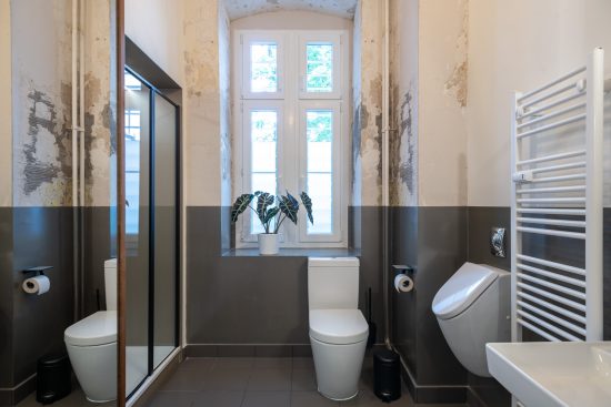 Barrierearmes Bad mit besonderem Charme: Ein präzise gespachtelter grau Wandsockel hebt sich von den unbehandelten Wänden im oberen Bereich ab. Neben dem Stand-WC ist Bewegungsfreiheit vorgesehen.