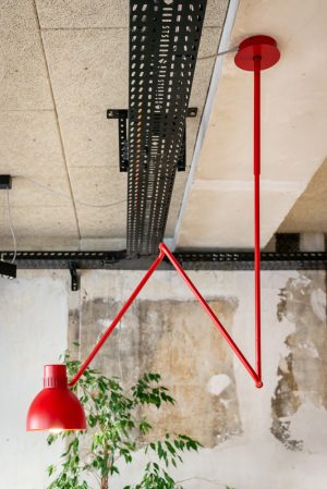 Detailaufnahme einer roten Deckenleuchte, schwarzer Elektrotrassen, der unbehandelt belassenen Wände und der Holzwolleplatten für die Raumakustik.