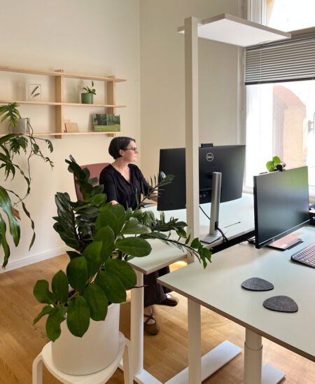 Heller Bürobereich mit Linoleum-Schreibtischen und Holzregal an einer weißen Wand. Eine Frau sitzt am Tisch und schaut aus dem Fenster. Pflanzen, Holz und freundliche Grüntöne erzeugen eine biophile Wohlfühlatmosphäre.