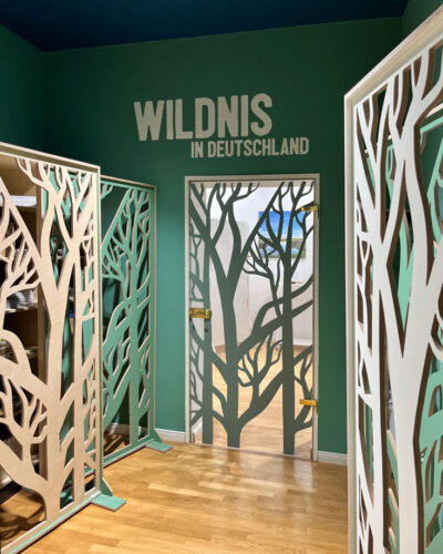Eingangsbereich mit dunkelgrünen Wänden, dunkelblauer Decke und Raumteilern mit Baumsilhouetten. An der Wand das Schrift-Logo "Wildnis in Deutschland".