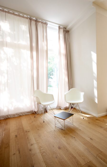 Zwei weiße Eames-Schalenstühle auf Eicheparkett vor einem Fenster mit Vorhang, durch de die Sonne scheint.