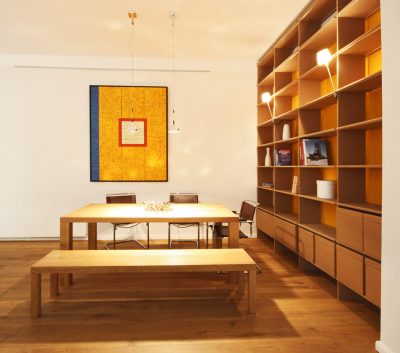 Essbereich mit Tisch und Sitzbank aus hellem Holz, rechts ein raumhohes Regal aus MDF mit oranger Rückwand. An der hinteren Wand ein großes abstraktes Bild in gelborange, blau und rot.