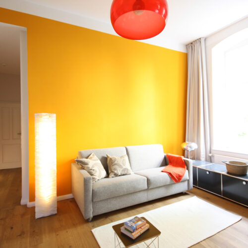 Sitzecke mit hellgrauem Sofa vor einer orangefarbenen Wand. Deckenleuchte aus rotem Kunststoff. Im Hintergrund ein USM-Haller-Lowboard in schwarz.